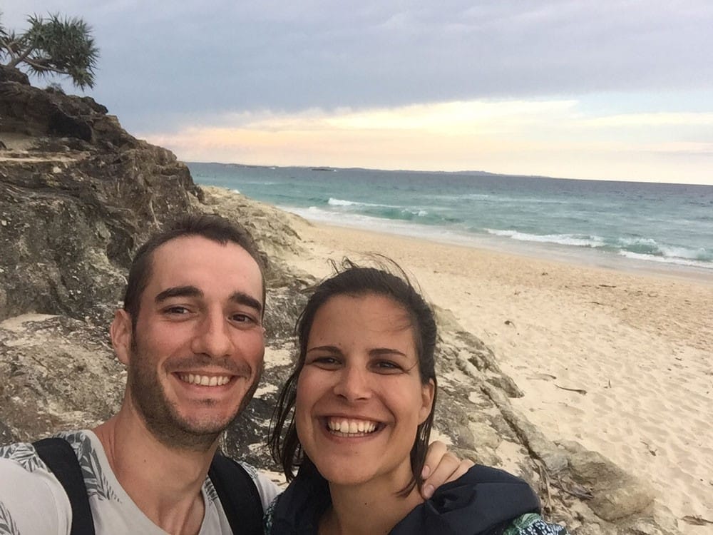 fotografía de un chico y una chica sonriendo a cámara y detrás la playa